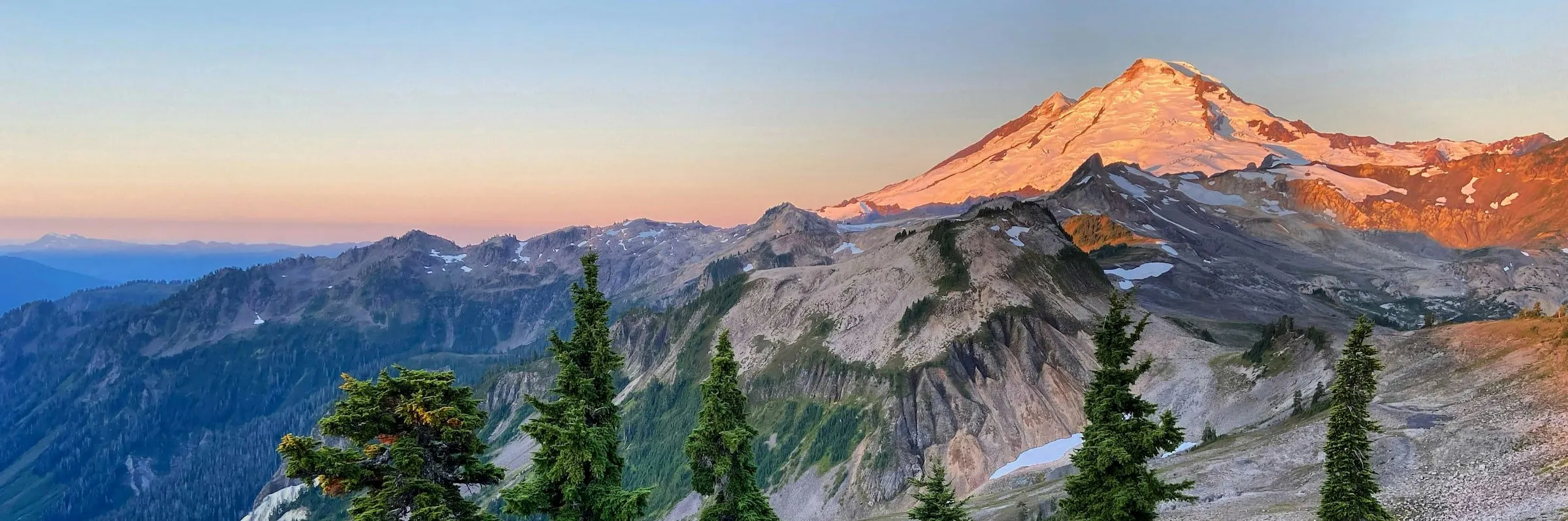 Sunset light on a snow-covered mountain beyond rocky ridges and evergreen trees.