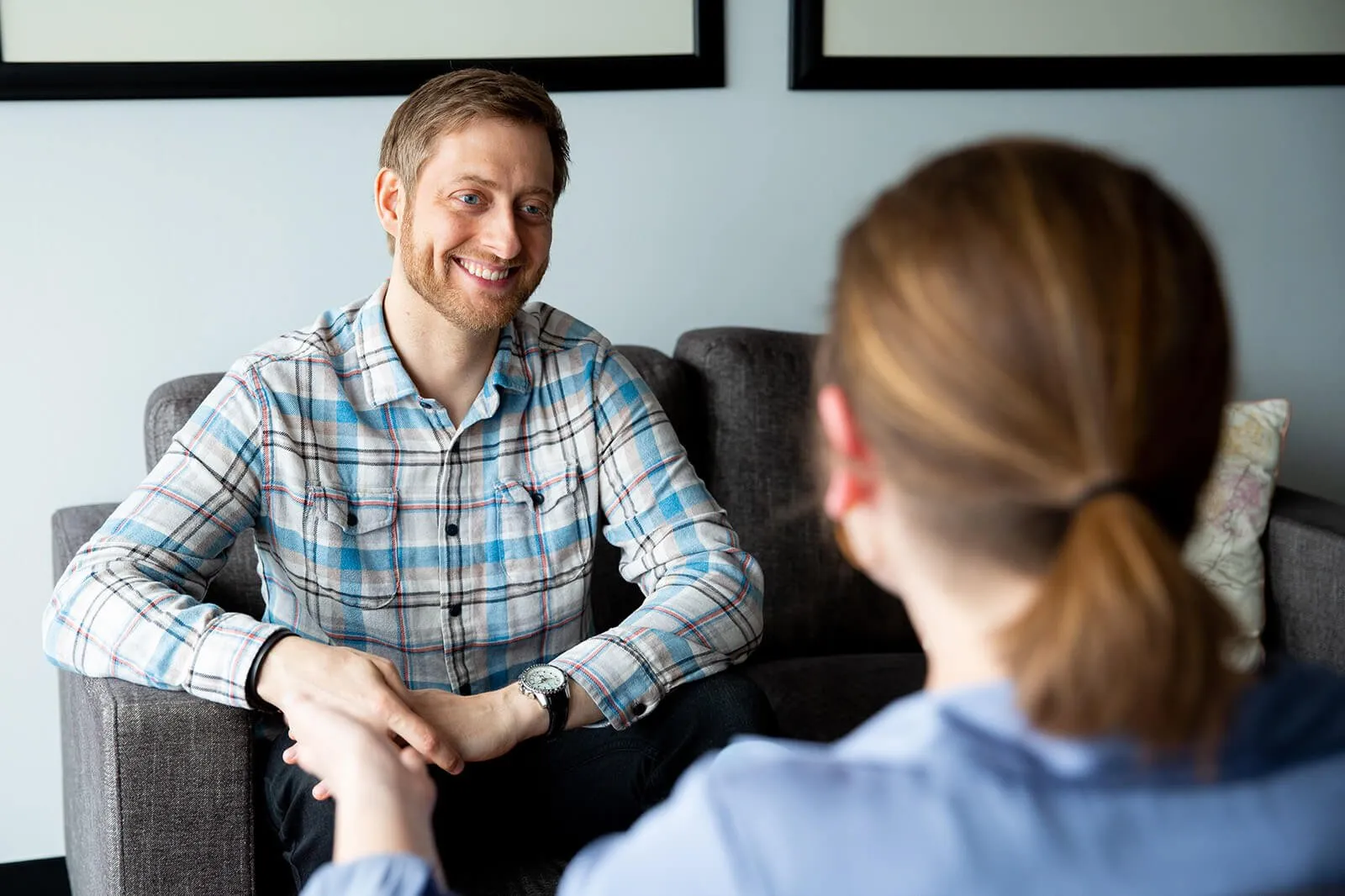 A man and woman engaged in a conversation, with the man smiling while sitting on a gray sofa in a modern room.