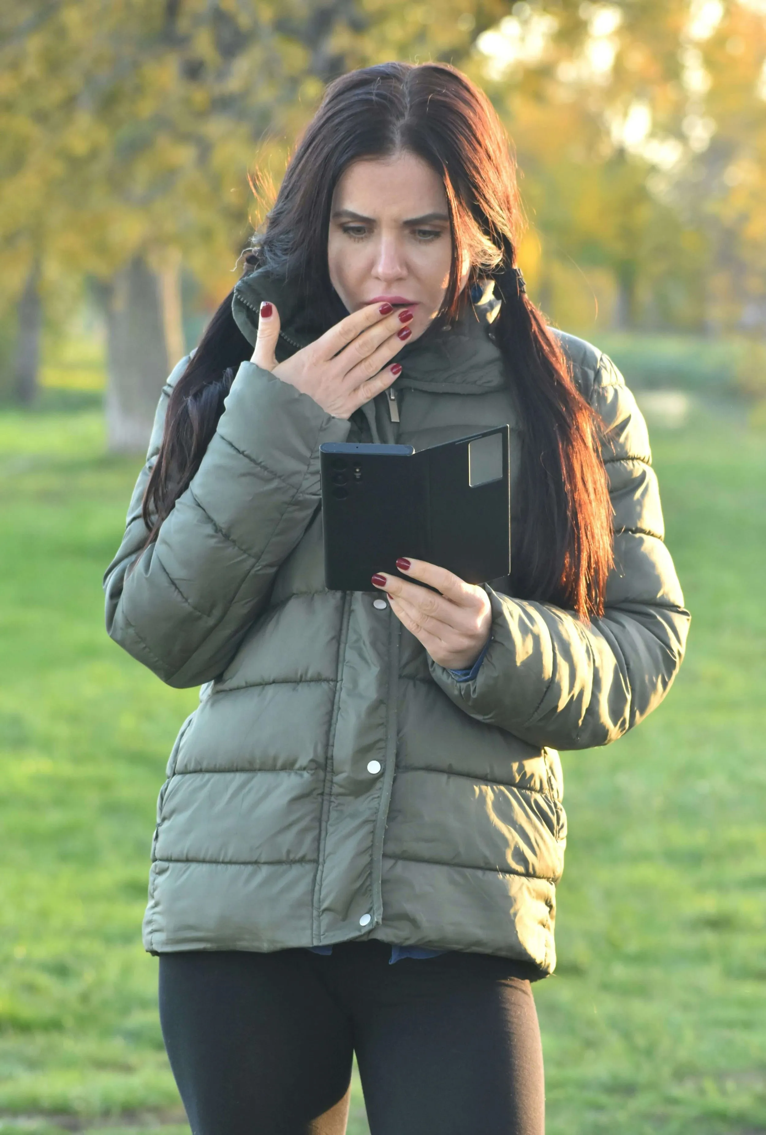 A person outdoors looking at a handheld device with one hand covering their mouth.