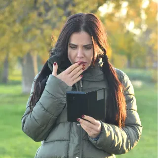 A person outdoors looking at a handheld device with one hand covering their mouth.