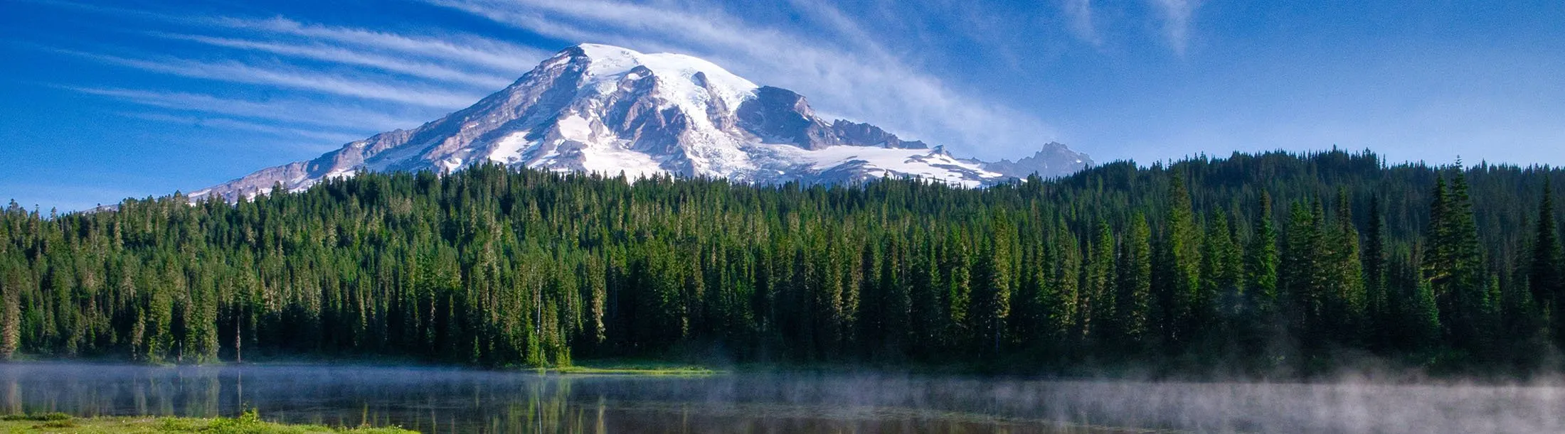 A snow-covered mountain above evergreen trees and a lake with reflections.